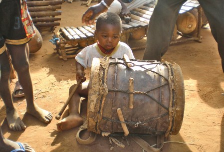 BURKIINA FASO 50 (EN UN POBLADO DE LA ETNIA SENUFO) TODO EL MUNDO PARTICIPA, ESTE MINUSCULO PERSONAJE CON UN TAMBOR CILINDRICO
